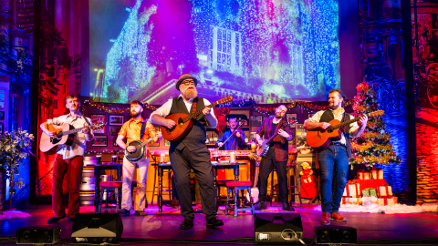 6 musicians stand on stage playing instrument, in front of a Christmassy background