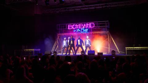 5 men perform on stage, a pink neon sign above them reads Boybands in the Buff