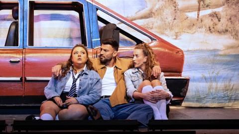 Three actors sat on stage in front of a red car.