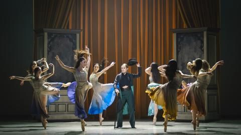 A production image of Northern Ballet's Gentleman Jack. A woman stands in the centre wearing a black suit, with a top hat, as other members dance around her wearing flowing dresses.