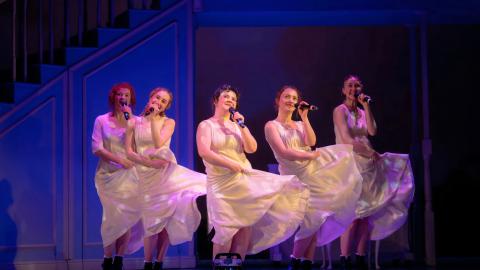 Five women in matching white dresses sing into microphones while stepping in unison on a stage