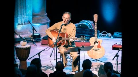 Musician Steve Craddock sits in a stage area of a cathedral playing guitar. Various instruments are set around him.