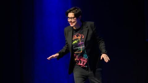 Sue Perkins stands on a dark stage in a black suit jacket and t-shirt, smiling at the audience