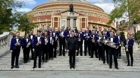 The Brighouse & Rastrick Band stand on the steps of a concert building, in suits, carrying brass instruments.