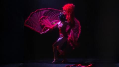 A woman in a long red cabaret skirt and feathers throws the skirt into the air, against a black background