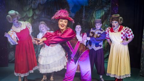 Lead performer in a bright pink costume poses at the front while a cast of colourful characters react behind on a storybook set.