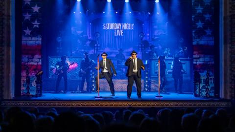 Two men in black suits perform on stage with blue lights shining on them.