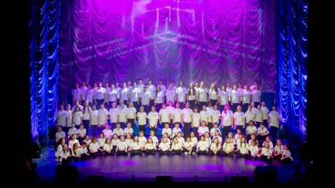 A large group of school children performing on stage against a purple background.