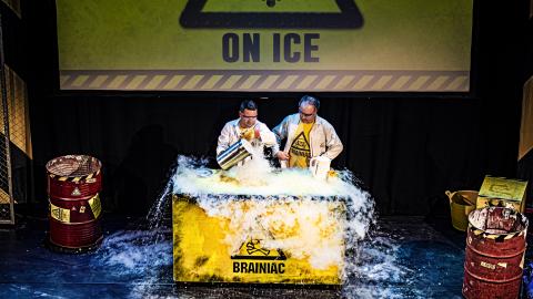 Two men wearing glasses and white lab coats stand on stage in front of a yellow podium with dry ice falling from it.