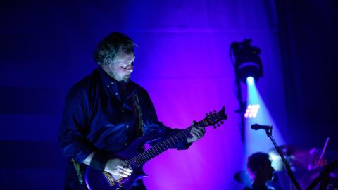 A man plays guitar on the left of the image, surrounded by purple stage lights.