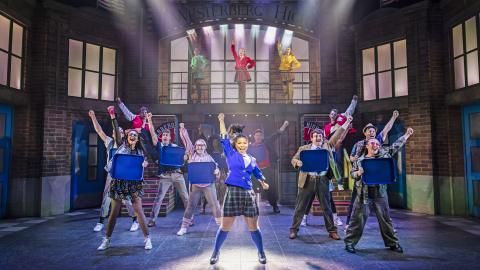 A group of people holding blue dinner trays punch their hands in the air on the set of Heathers The Musical.