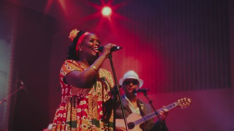 A woman wearing a long yellow dress sings into a microphone with a man playing guitar behind her and red lights shining onto the stage.