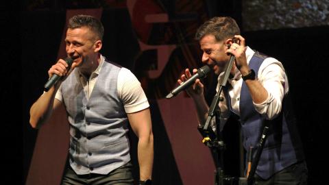 Two musicians perform on stage, singing into microphones. They both wear white shirts and smart waistcoats.