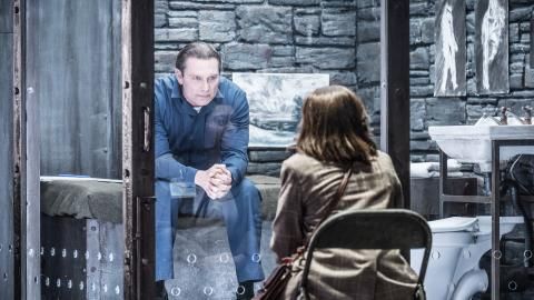 A production image for Silence of the Lambs, a man sits in a cell staring at a woman sat on the other side of some glass.