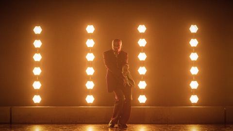 A man dances on stage surrounded by orange light.