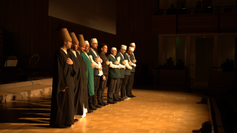 A group of men stand on stage wearing long outfits and holding their hand to their chest.
