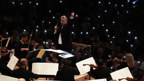 A man in a black suit leads an orchestra on stage, reading sheet music