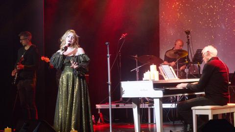 A woman sings into a microphone on stage wearing a long, gold dress. A man plays piano on the right.