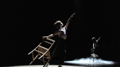 A young boy dances holding a chair on stage, against a black background.