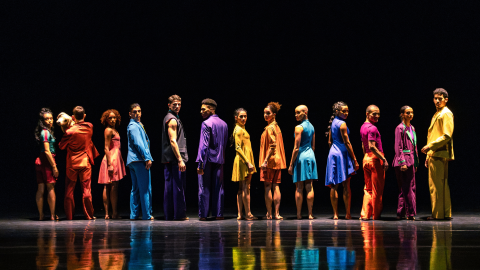 A line of dancers all wearing bright, different coloured outfits against a black background. 