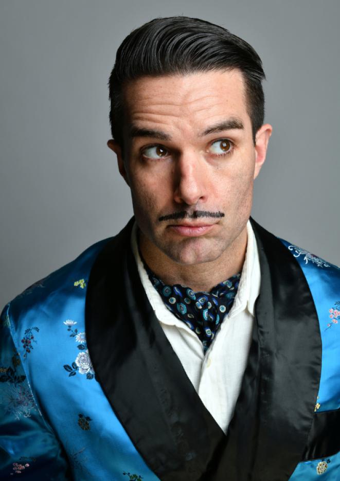Comedian Troy Hawke stands in front of a grey background, wearing a smart blue and black jacket.