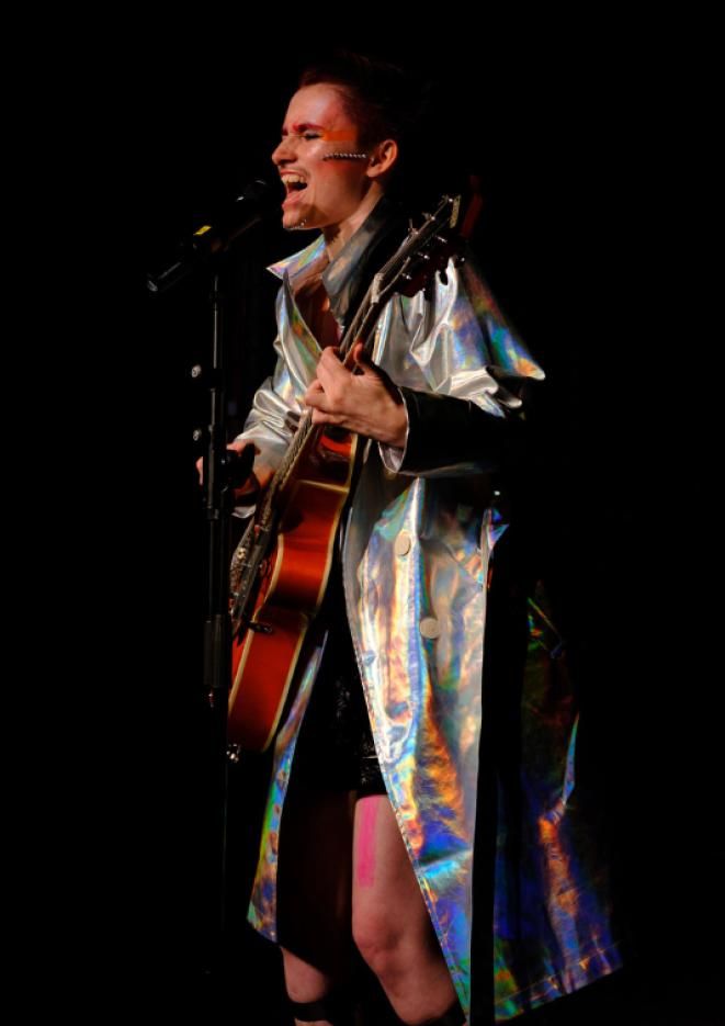 Drag performer stands on stage playing guitar and singing into a microphone.