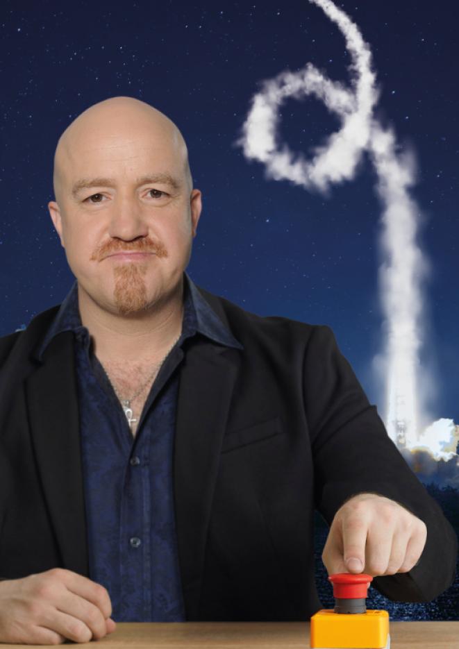 Comedian Andy Parsons sits at a desk pushing a button.