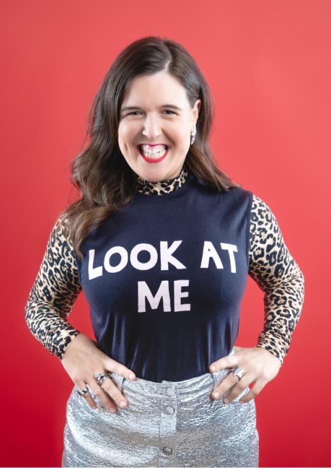 Comedian Rosie Jones stood against a bright red background with a black tshirt with white text that reads Look At Me.