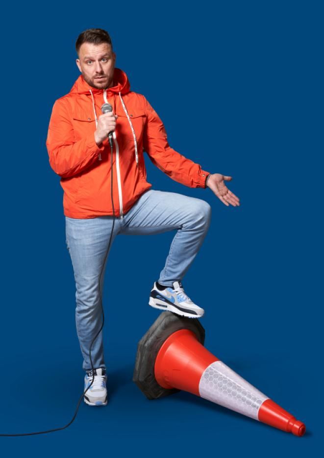 Comedian Daniel O'Reilly stood against a dark blue background with one foot resting on a traffic cone.
