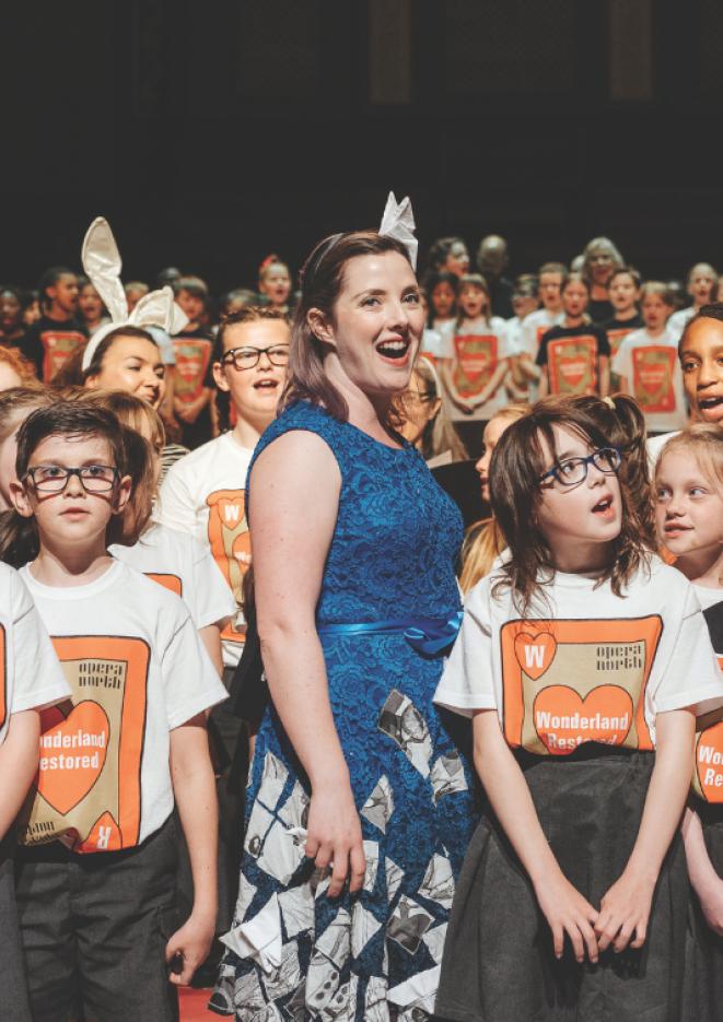 A woman stood in the centre of the image wearing a blue dress, with young students around her wearing white and orange t-shirts.
