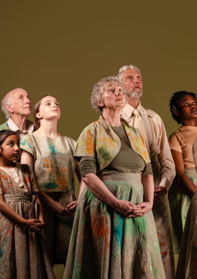 A group of performers stood against a plain background wearing earth tones and looking up.