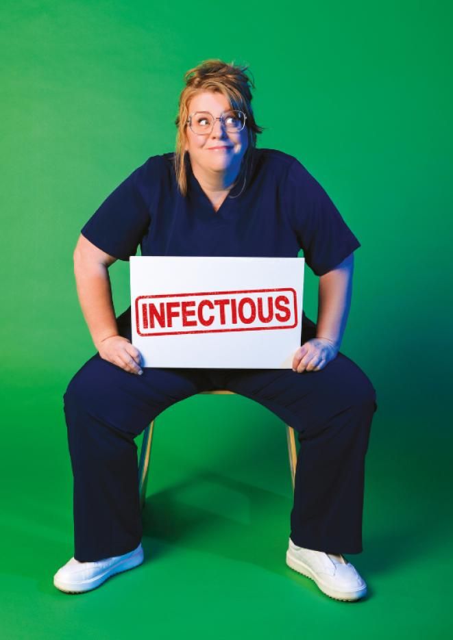 Comedian Georgie Caroll sat on a stool against a green background holding a white sign that reads Infectious.