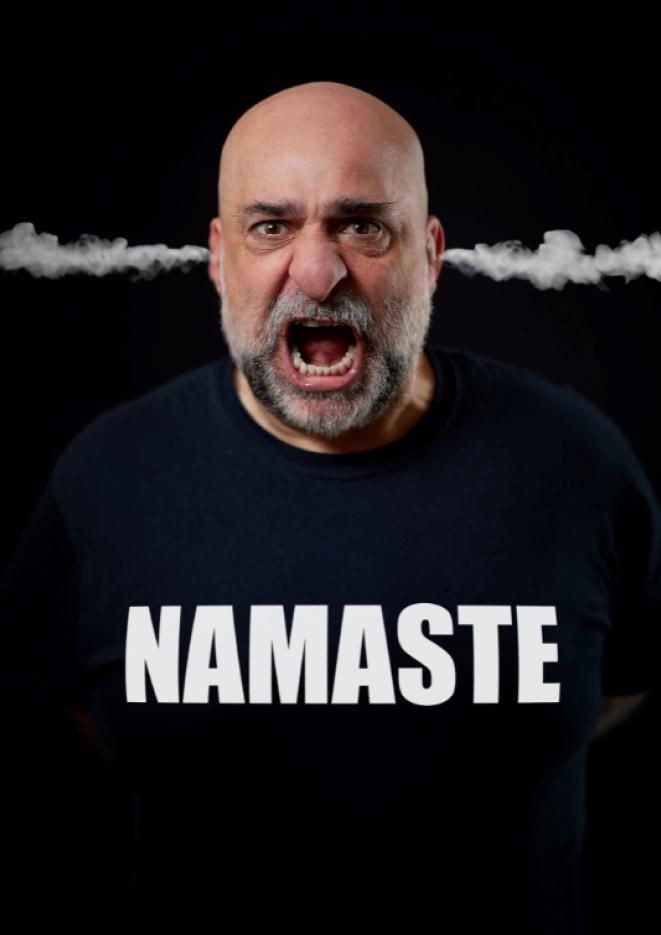 Comedian Omid Djalili stands against a black background wearing a black jumper that reads Namaste.