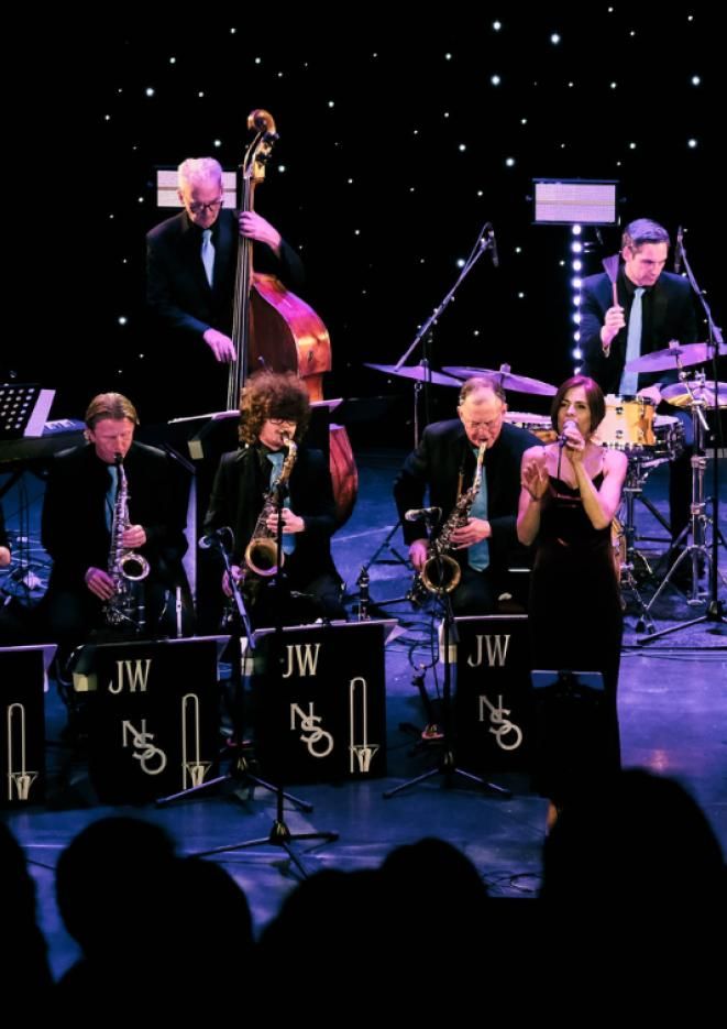 An orchestra of musicians sat against a black starry background playing instruments, with a woman stood singing into a microphone