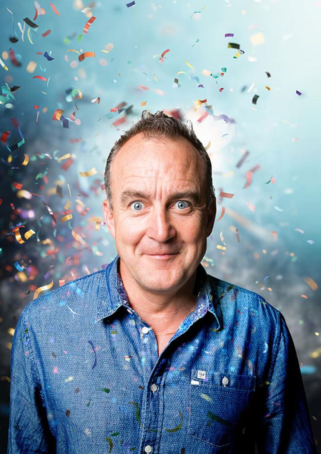 Portrait of comedian Jimeoin wearing a blue shirt, with confetti covering him.