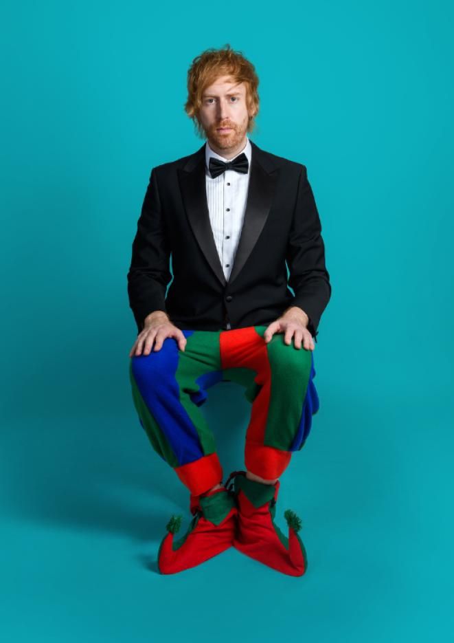 Comedian Mark Simmons sits against a plain, teal backdrop, wearing jester pants and shoes.