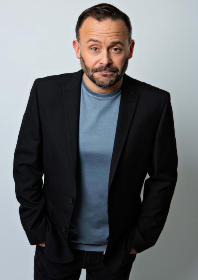 Portrait of comedian Geoff Norcott stands against a plain, grey background wearing a blue T-Shirt and black blazer.