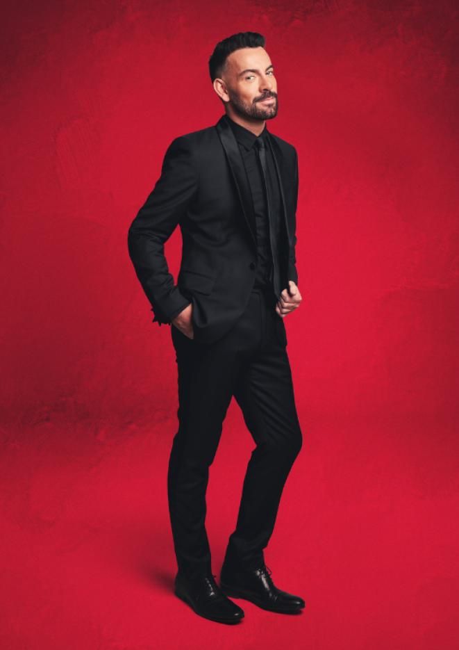 Singer and actor Ben Forster wears an all black suit, stood in front of a red background.
