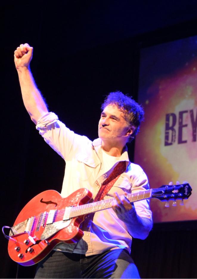 TV Personality and vet Noel Fitzpatrick stands on stage playing guitar and raising his arm in the air.
