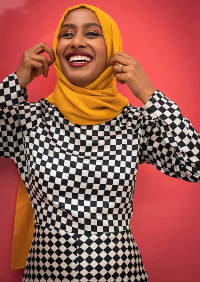 Comedian Ola Labib stands against a bright pink background smiling.