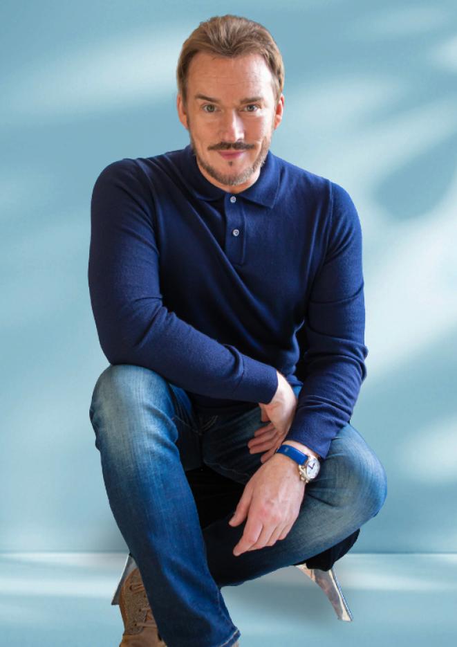 Singer Russell Watson sits on a silver stool against a blue background.