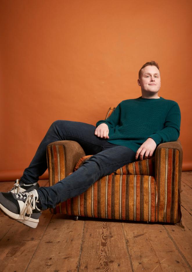 Comedian Josh Jones lounges on an orange, stripey seat against a plain orange background.