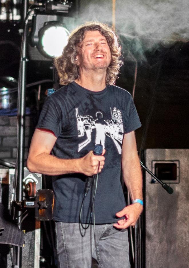 Danny McNamara in a black t-shirt, holding a microphone, smiling on stage
