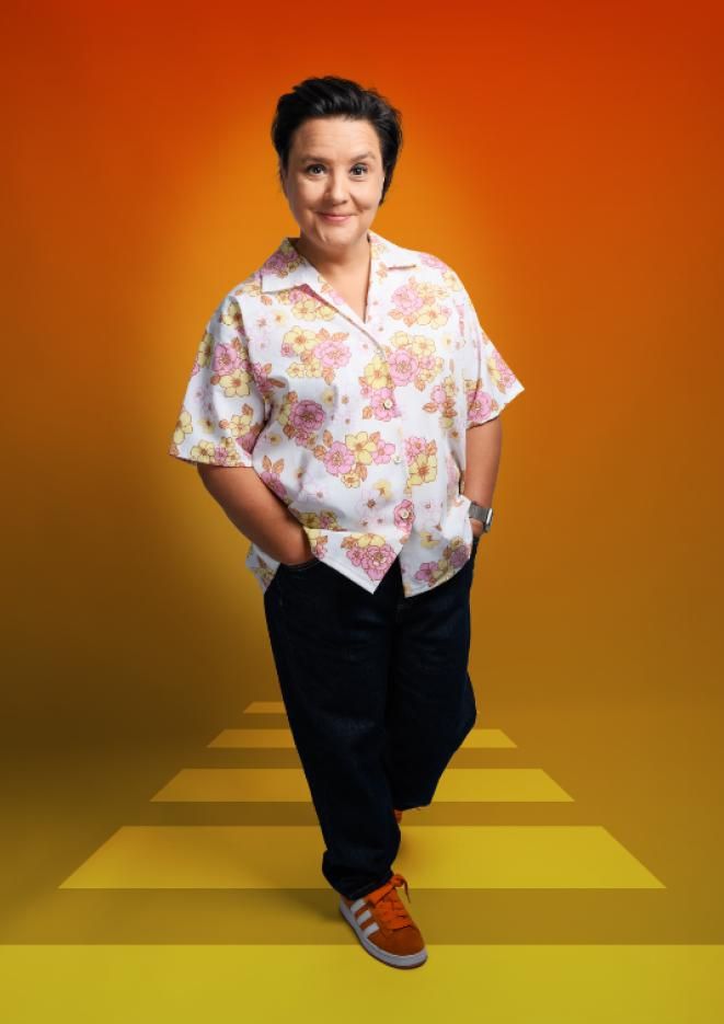 Susan Calman wearing a white and orange shirt, walking to the camera in front of an orange background