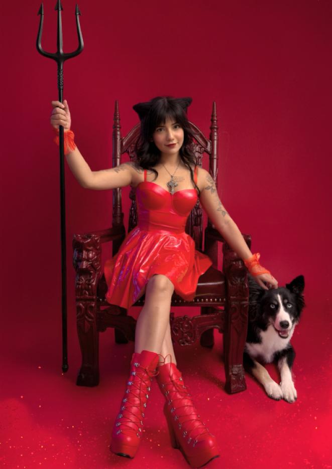 A woman sits on a chair against a red background with a collie dog at her side.