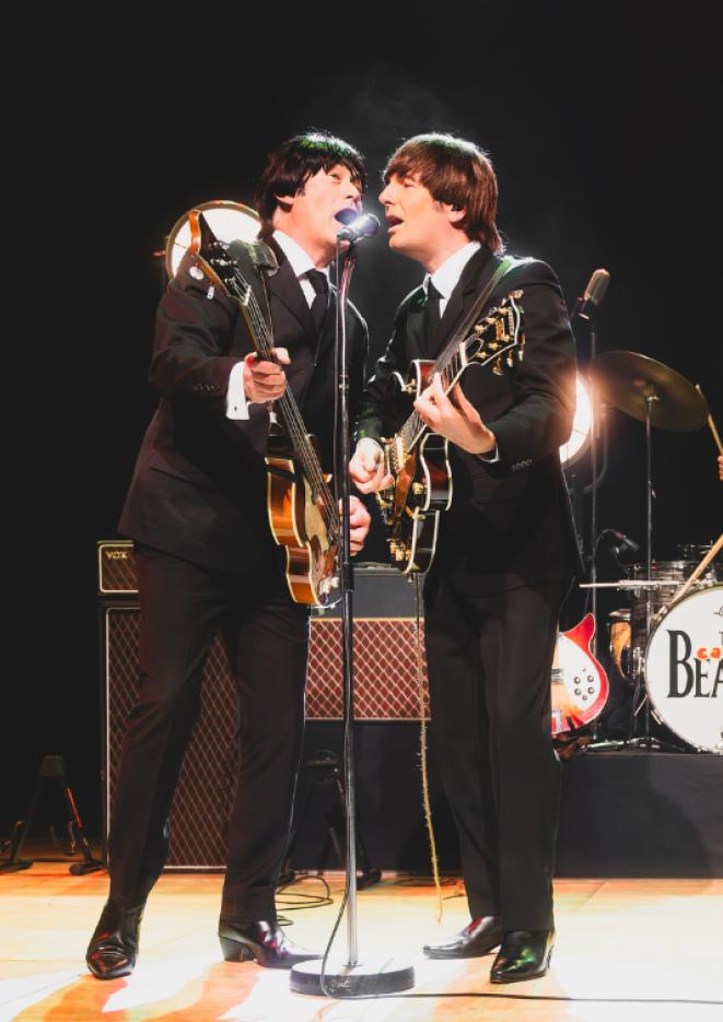 Two men dressed in suits singing into a microphone, playing the guitar