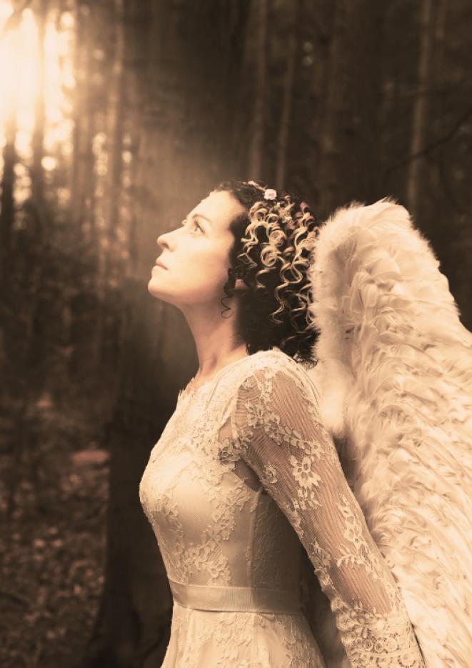 Kate Rusby in a white lacy dress, with large white angel wings on her back, looking up