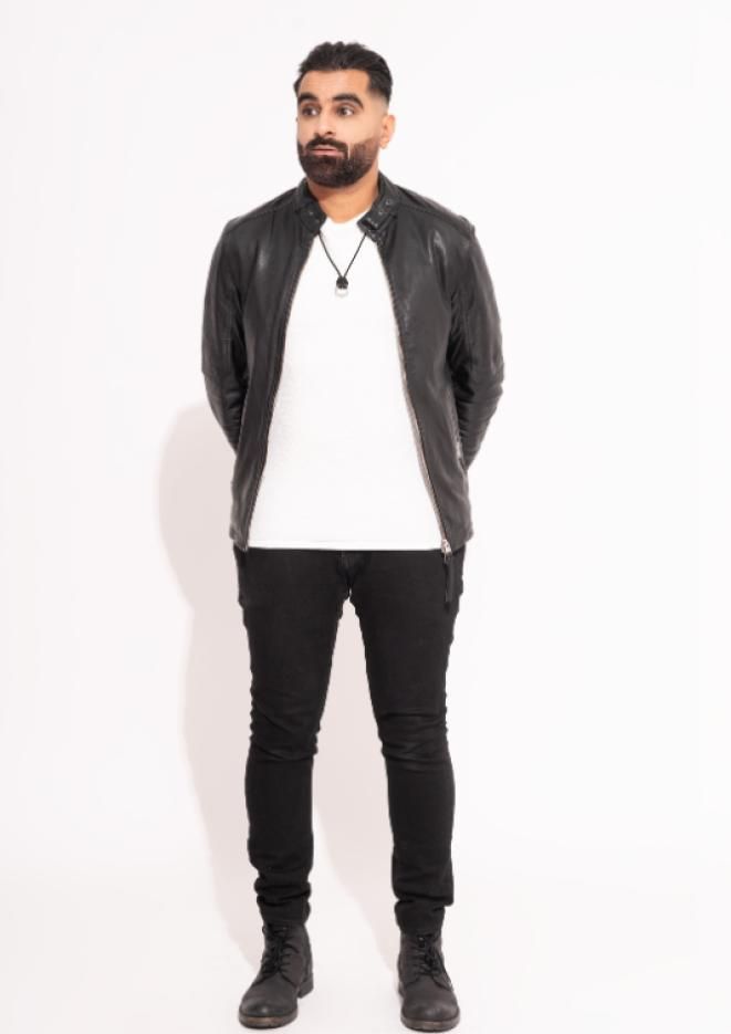 Comedian Tez Ilyas stands against a plain, white background, wearing a white t-shirt, black jeans and jacket. 