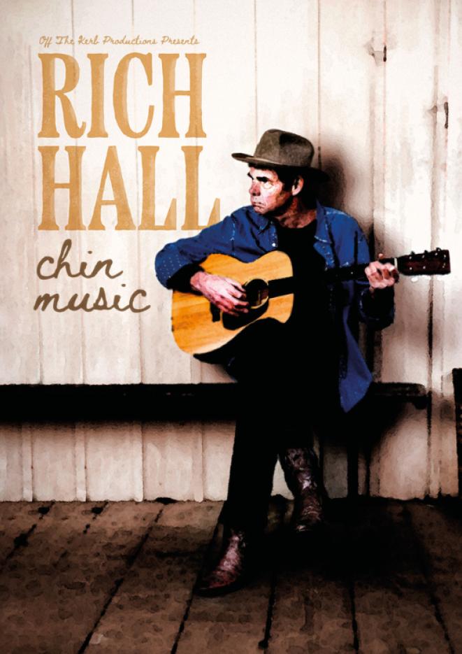 Comedian Rich Hall sits against a wood wall holding a guitar.