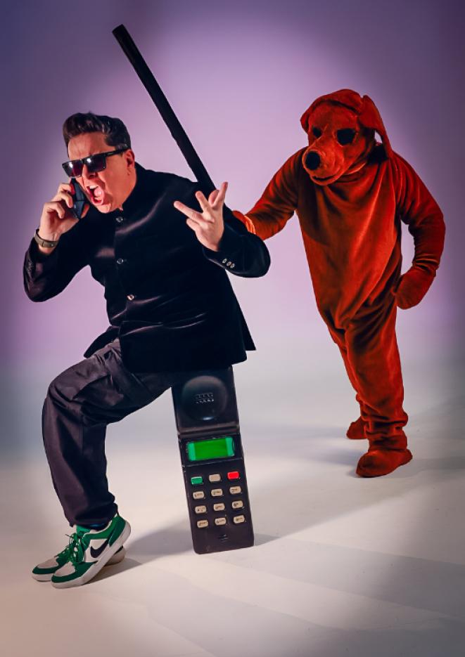 Comedian Dom Joly sits on a giant phone against a purple background, with a man in a red dog outfit stood behind him.